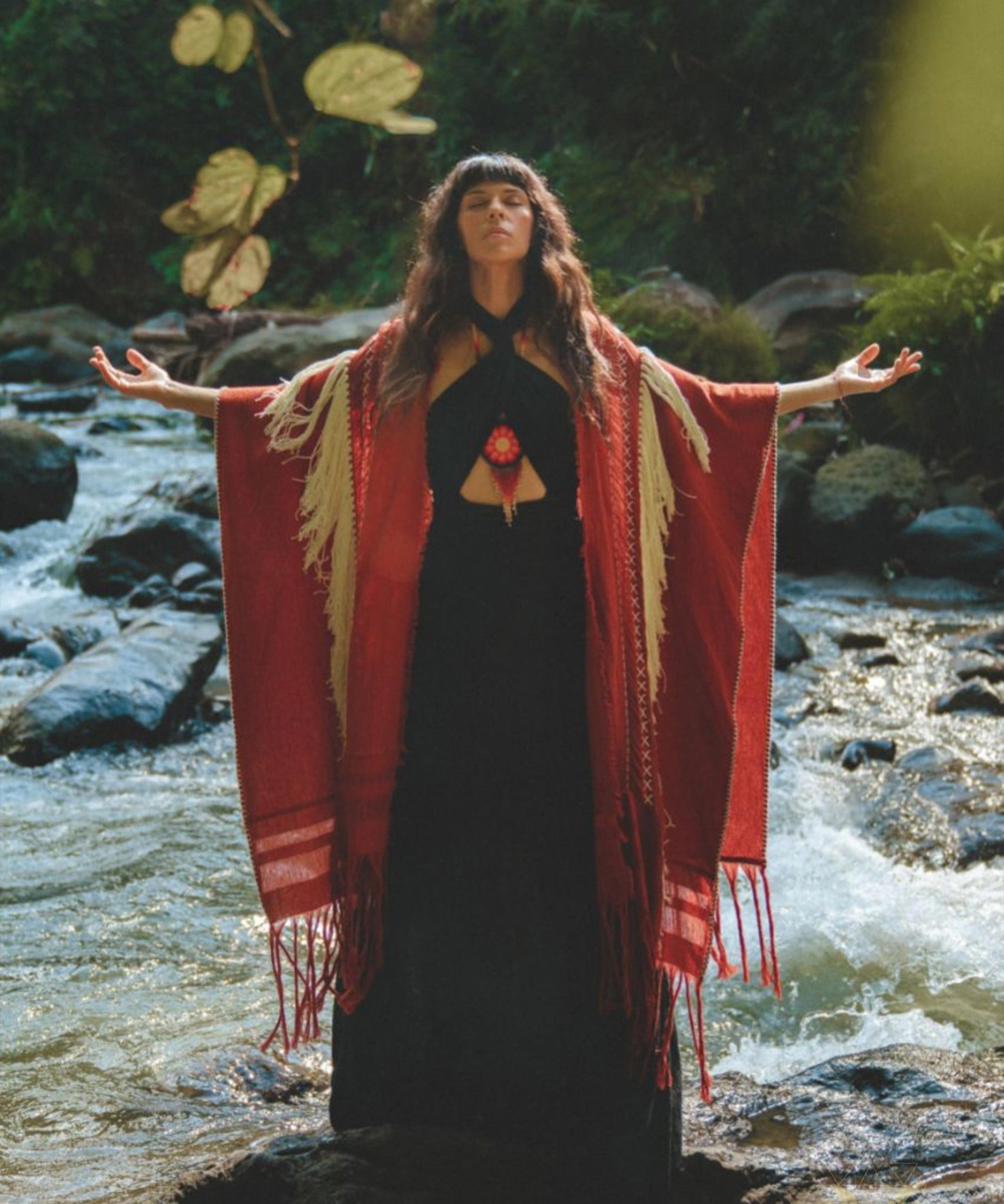 A woman stands on rocks in a stream, arms spread wide and looking upward. She is wearing the Black Harmonia Dress from AYA Sacred Wear, a Gothic sexy, adjustable dress also known as the Fairy of the River Dress. The dress is crafted from organic linen and cotton and is complemented by a red and beige shawl. The surrounding area is lush with greenery as sunlight filters through the trees.