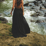 A woman with long, wavy hair, dressed in a flowing Black Harmonia Dress from AYA Sacred Wear—an adjustable Gothic sexy dress known as the Fairy of the River Dress and made from organic linen and cotton dyed with botanical dyes—stands on a rocky ledge overlooking a fast-moving stream surrounded by rocks and greenery. The scene exudes a sense of peace and natural beauty.
