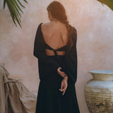 A woman with long hair is seen from behind, wearing a black tank top and maxi skirt from AYA Sacred Wear. The outfit, made of lightweight cotton fabric, features an open back with cross straps. She stands with her arms positioned behind her, surrounded by large clay pots and draped fabric, under the shade of palm leaves.