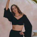 A woman with long hair confidently poses in a Black Summer Open Belly Top and Skirt from AYA Sacred Wear. The black, two-piece outfit made from lightweight cotton fabric stands out against a soft pink background. Beneath large green palm leaves, she rests one hand on the leaf while the other is placed on her hip, crafting a tropical and stylish atmosphere.