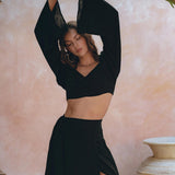 A woman in an elegant AYA Sacred Wear ensemble, featuring the Black Summer Open Belly Top and Skirt, poses gracefully with her arms raised against a textured pastel background. She stands next to a large decorative vase, surrounded by tropical leaves, exuding elegance and style.
