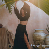 A woman dressed in the Black Summer Open Belly Top and Skirt ensemble from AYA Sacred Wear stands with arms raised, gracefully holding palm leaves. She poses barefoot on a stone floor amidst large pottery and soft drapery, bathed in streaming sunlight that highlights her chic outfit.