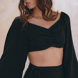 A woman with long brown hair is wearing an elegant Black Summer Open Belly Top and Skirt from AYA Sacred Wear, crafted with lightweight cotton fabric and featuring ruched detailing with long sleeves. She stands against a light, soft-focus background, looking down to her left with a serene expression.