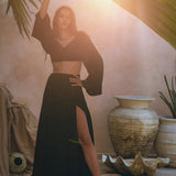 A woman models the Black Summer Open Belly Top and Skirt by AYA Sacred Wear, posing gracefully under a tropical palm frond. She stands barefoot on a sandy floor, encircled by large pottery and a shell-shaped sculpture. Sunlight filters through the lightweight cotton fabric of her black tank top and maxi skirt, creating a warm, serene atmosphere.
