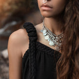 A young woman with long wavy hair stands outdoors, gazing to the side. She is wearing an AYA Sacred Wear Black Aurora Greek Goddess Dress — a Cold Shoulder Maxi Dress with gothic Grecian flair. Her look is accessorized with multiple silver coin necklaces and a nose ring. The blurred background, softly lit by warm sunlight, creates a natural ambiance.