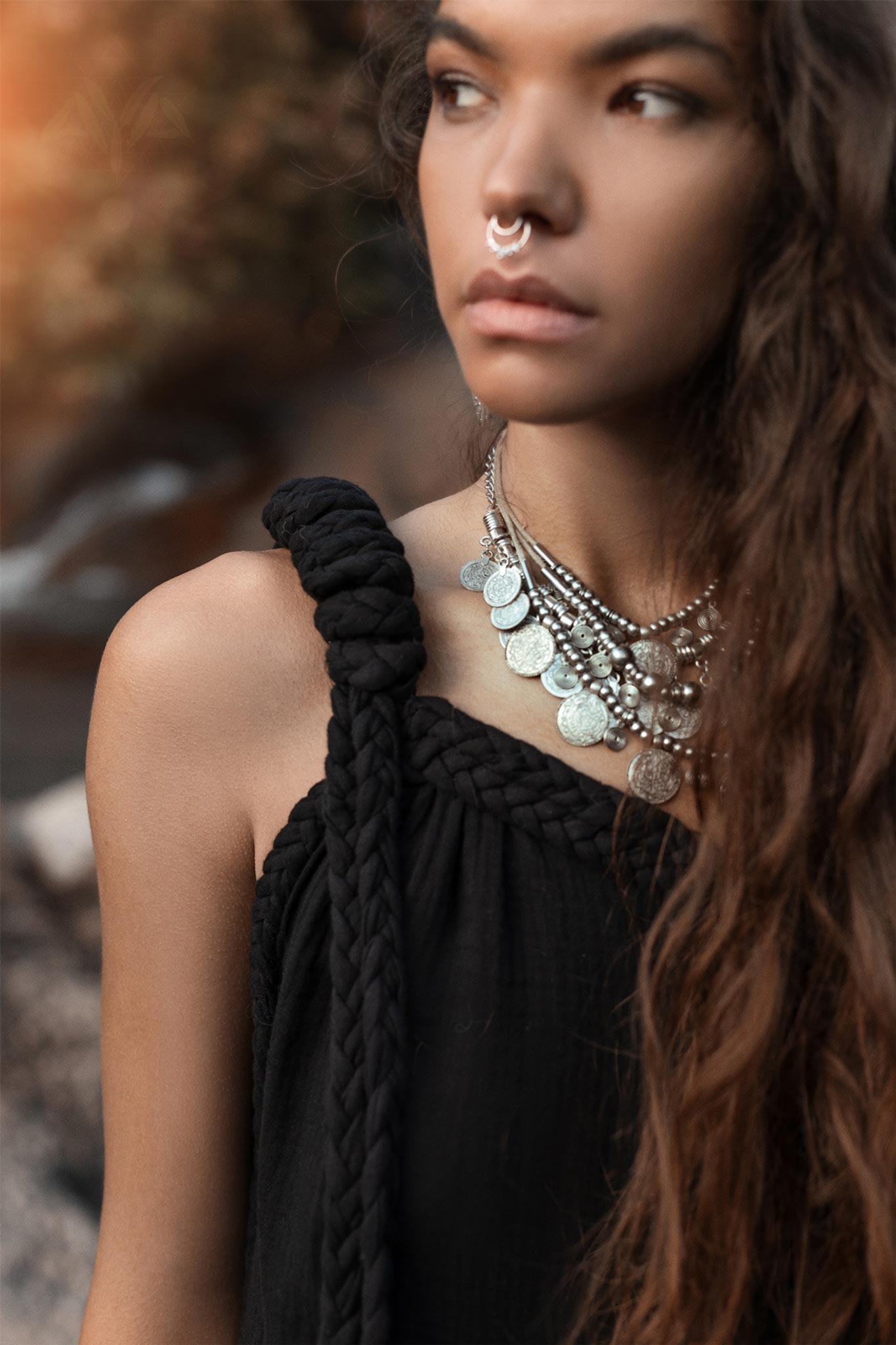 A young woman with long wavy hair stands outdoors, gazing to the side. She is wearing an AYA Sacred Wear Black Aurora Greek Goddess Dress — a Cold Shoulder Maxi Dress with gothic Grecian flair. Her look is accessorized with multiple silver coin necklaces and a nose ring. The blurred background, softly lit by warm sunlight, creates a natural ambiance.