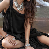 A woman with long wavy hair sits on a rock beside a river, wearing the Black Aurora Greek Goddess Dress by AYA Sacred Wear. The cold shoulder maxi dress, adorned with rope straps, complements her various silver jewelry including a layered necklace, earrings, bangles, and rings. She gazes downward with her legs crossed.