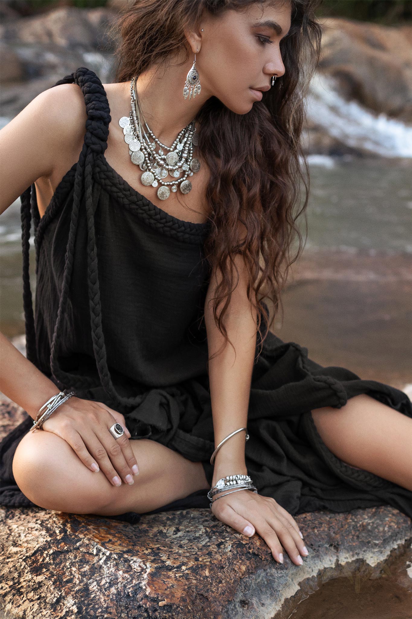 A woman with long wavy hair sits on a rock beside a river, wearing the Black Aurora Greek Goddess Dress by AYA Sacred Wear. The cold shoulder maxi dress, adorned with rope straps, complements her various silver jewelry including a layered necklace, earrings, bangles, and rings. She gazes downward with her legs crossed.