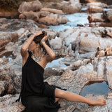 A woman in an AYA Sacred Wear Black Aurora Greek Goddess Dress • Cold Shoulder Maxi Dress • Gothic Grecian Dress sits barefoot on rocky terrain near a flowing river. She is adorned with several bracelets and a large necklace. One arm is raised to adjust her hair, and her other arm rests on the ground. The background features trees and water.