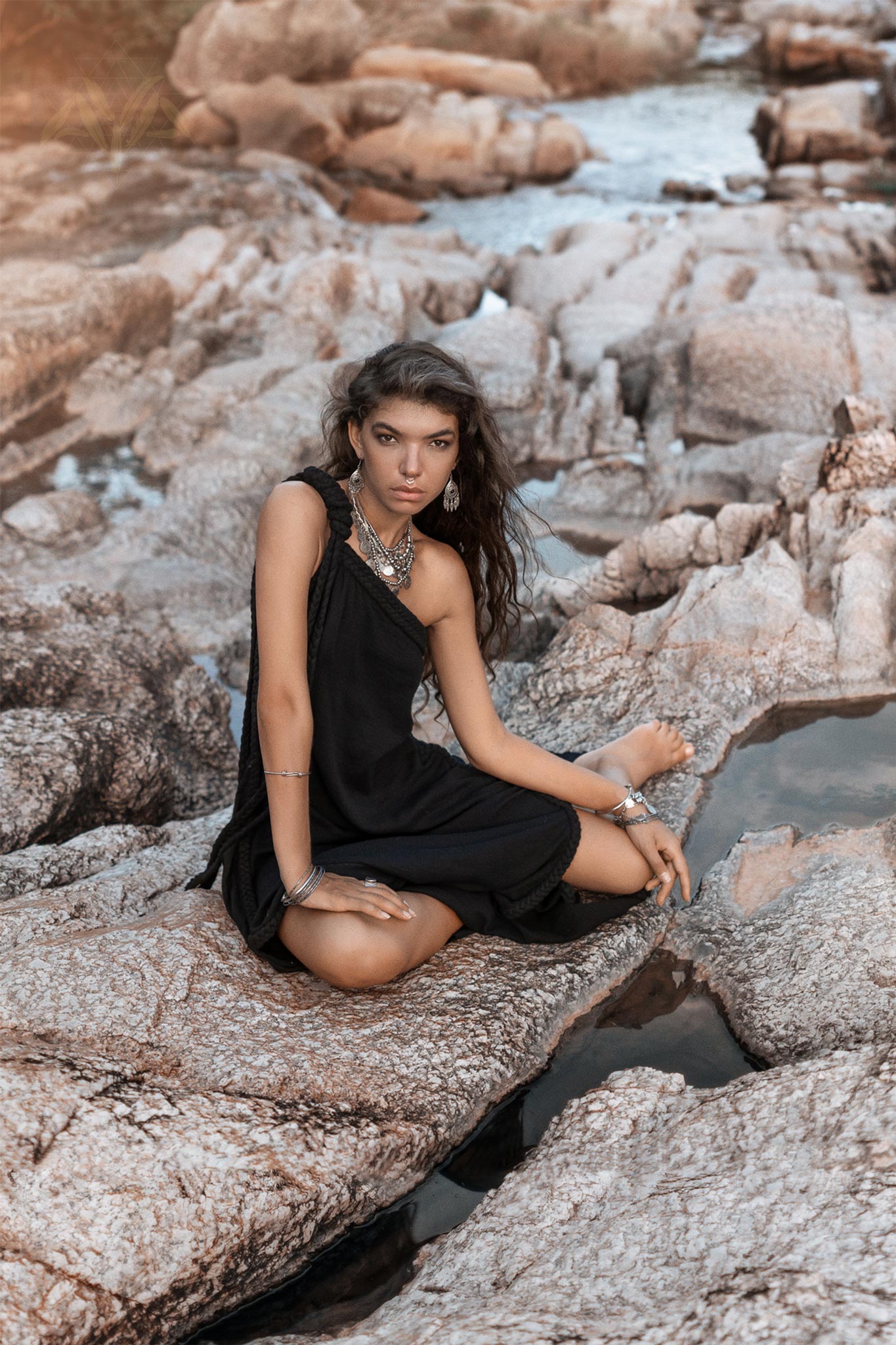 A woman wearing the Black Aurora Greek Goddess Dress by AYA Sacred Wear, crafted from organic cotton, sits on rocky terrain near a small body of water. She has long, wavy brown hair and is adorned with multiple pieces of jewelry. She looks directly at the camera with a calm expression while one leg is crossed over the other.