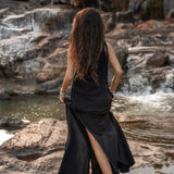 A woman with long, wavy hair walks barefoot on rocky terrain beside a stream. She is wearing the Black Aurora Greek Goddess Dress by AYA Sacred Wear, a cold shoulder maxi dress made of organic cotton, featuring a high slit. The background includes a rocky landscape with water flowing over the stones and trees with autumn foliage.