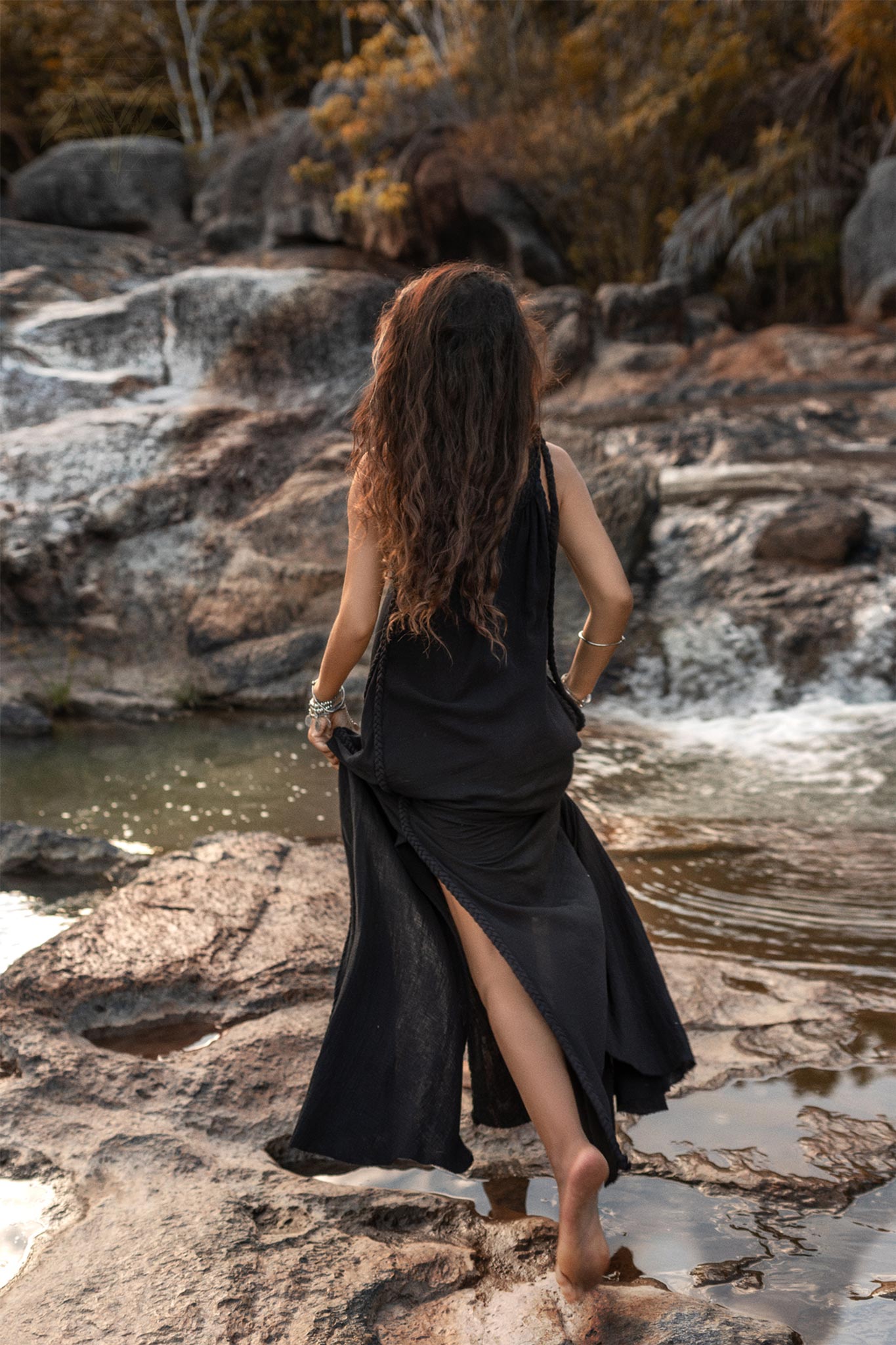 A woman with long, wavy hair walks barefoot on rocky terrain beside a stream. She is wearing the Black Aurora Greek Goddess Dress by AYA Sacred Wear, a cold shoulder maxi dress made of organic cotton, featuring a high slit. The background includes a rocky landscape with water flowing over the stones and trees with autumn foliage.