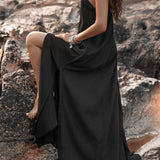 A woman in an AYA Sacred Wear Black Aurora Greek Goddess Dress – a Gothic Grecian-style cold shoulder maxi dress – sits on a rocky landscape. Her hair flows loosely, and she accessorizes with bracelets and necklaces. The scene has a serene, natural backdrop featuring water and boulders. Her pose is relaxed, with one knee bent and her arms resting gently.