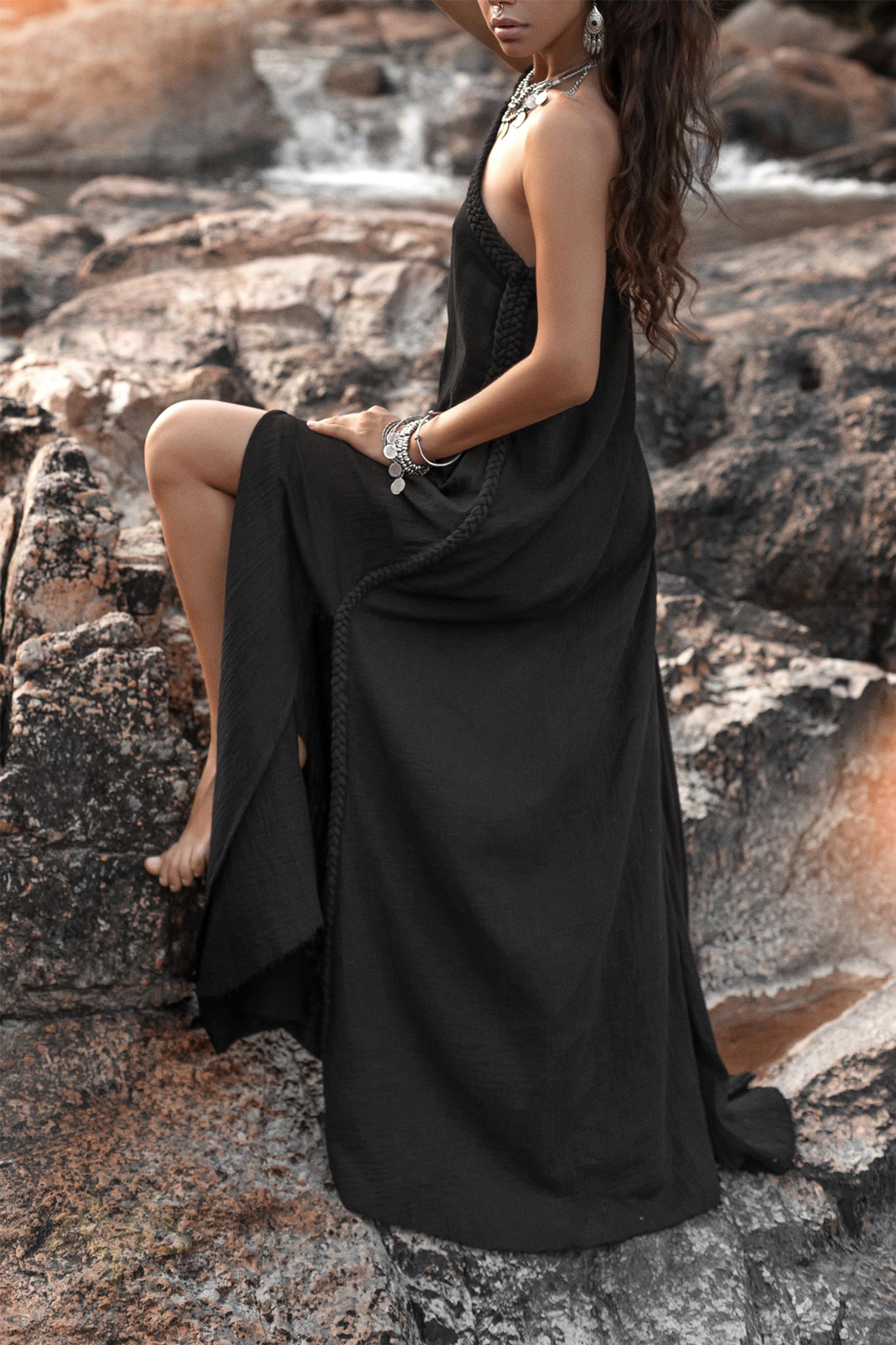 A woman in an AYA Sacred Wear Black Aurora Greek Goddess Dress – a Gothic Grecian-style cold shoulder maxi dress – sits on a rocky landscape. Her hair flows loosely, and she accessorizes with bracelets and necklaces. The scene has a serene, natural backdrop featuring water and boulders. Her pose is relaxed, with one knee bent and her arms resting gently.