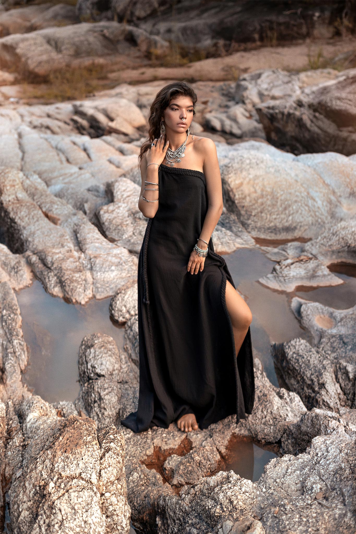 A woman wearing a long, Cold Shoulder Black Aurora Greek Goddess Maxi Dress by AYA Sacred Wear, adorned with various pieces of jewelry, stands barefoot on a rocky terrain. She gazes thoughtfully into the distance, her hand resting on her face, as the slit in her Gothic Grecian dress reveals part of her leg. The background features rugged rocks.