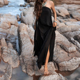 A woman with long, wavy hair wears a flowing Black Aurora Greek Goddess Dress from AYA Sacred Wear. She is adorned with ornate jewelry and walks barefoot on rocky terrain beside a stream. The Cold Shoulder Maxi Dress, also known as the Gothic Grecian Dress, features a high slit that exposes one leg. The backdrop of rocks and water creates a rugged, natural setting.