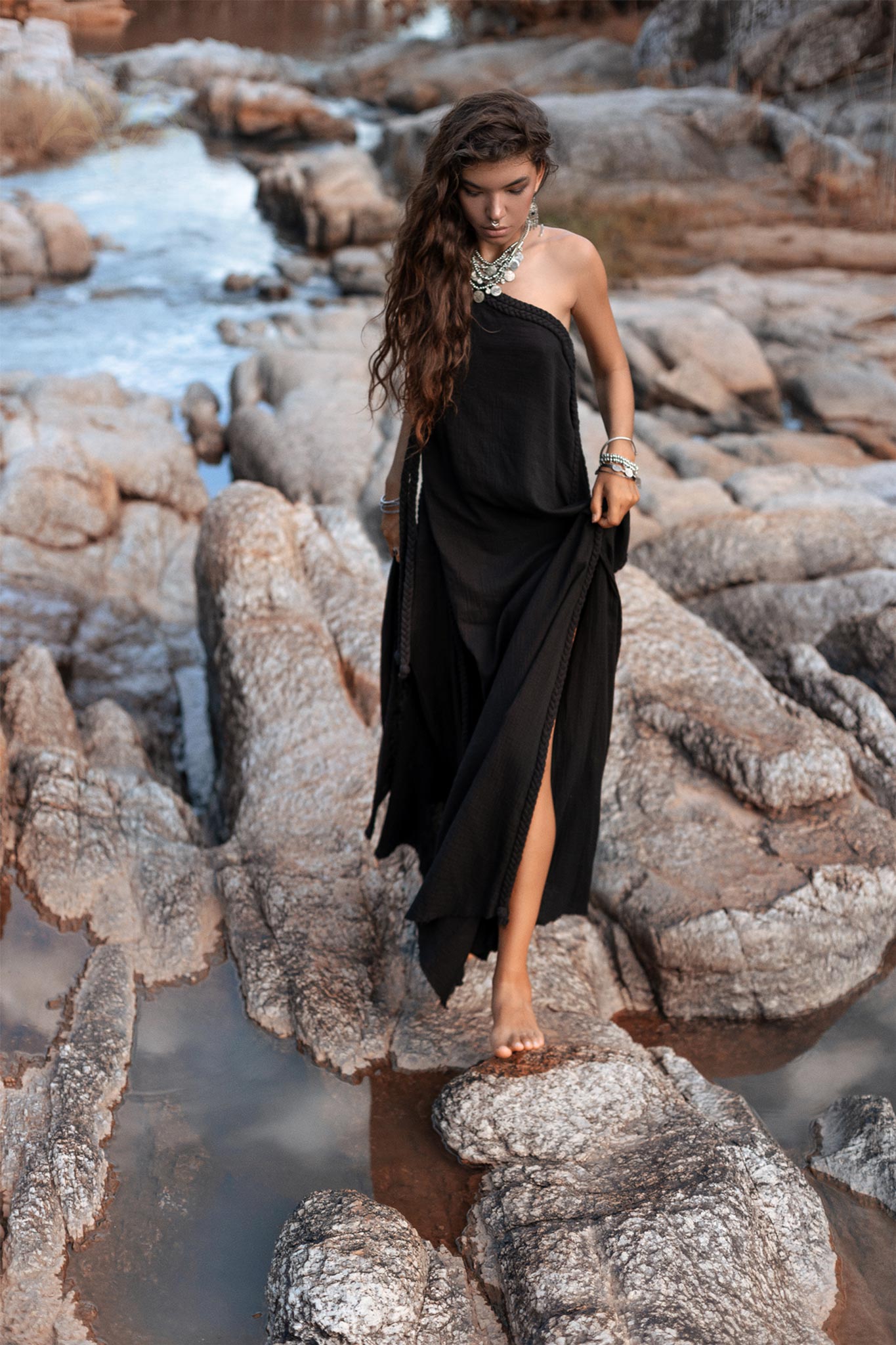 A woman with long, wavy hair wears a flowing Black Aurora Greek Goddess Dress from AYA Sacred Wear. She is adorned with ornate jewelry and walks barefoot on rocky terrain beside a stream. The Cold Shoulder Maxi Dress, also known as the Gothic Grecian Dress, features a high slit that exposes one leg. The backdrop of rocks and water creates a rugged, natural setting.