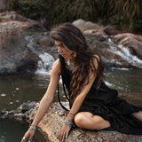 A woman with long, wavy hair sits on a rock by the water's edge, dressed in a flowing Black Aurora Greek Goddess Dress - Cold Shoulder Maxi Dress • Gothic Grecian Dress by AYA Sacred Wear, adorned with several bracelets and jewelry. She extends one hand to touch the surface of the water, surrounded by a natural, wooded backdrop.