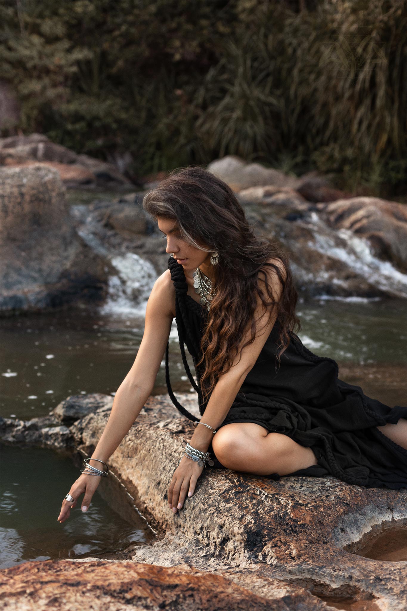 A woman with long, wavy hair sits on a rock by the water's edge, dressed in a flowing Black Aurora Greek Goddess Dress - Cold Shoulder Maxi Dress • Gothic Grecian Dress by AYA Sacred Wear, adorned with several bracelets and jewelry. She extends one hand to touch the surface of the water, surrounded by a natural, wooded backdrop.