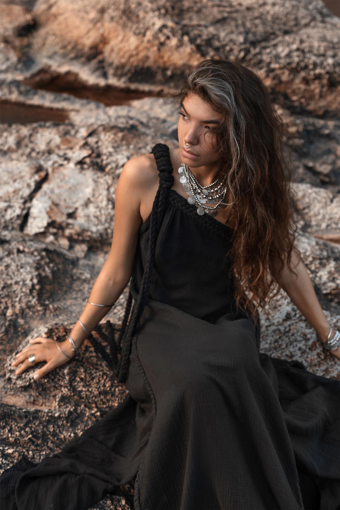 A woman with long, wavy hair sits on a rocky surface, wearing the Black Aurora Greek Goddess Dress by AYA Sacred Wear. The Cold Shoulder Maxi Dress and multiple layered necklaces complement her look. With a contemplative expression, she gazes to the side against a backdrop featuring rough, textured rocks.