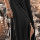 An individual dressed in a luxurious, flowing Black Aurora Greek Goddess Dress from AYA Sacred Wear stands on rugged terrain. This Cold Shoulder Maxi Dress, also known as the Gothic Grecian Dress, features intricate braided details and a slit that elegantly reveals part of the leg. With silver bangles and a ring adorning their right hand, their hair gracefully falls to one side.