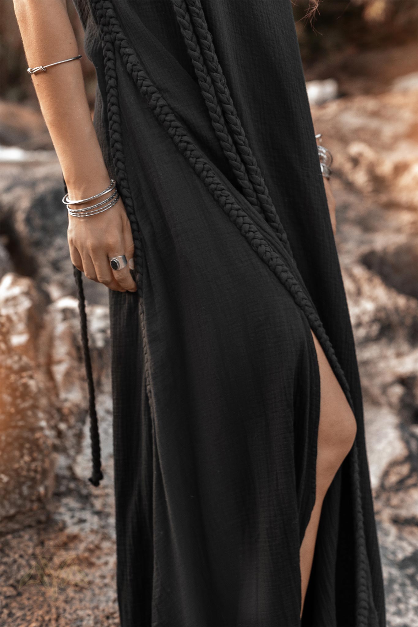 An individual dressed in a luxurious, flowing Black Aurora Greek Goddess Dress from AYA Sacred Wear stands on rugged terrain. This Cold Shoulder Maxi Dress, also known as the Gothic Grecian Dress, features intricate braided details and a slit that elegantly reveals part of the leg. With silver bangles and a ring adorning their right hand, their hair gracefully falls to one side.