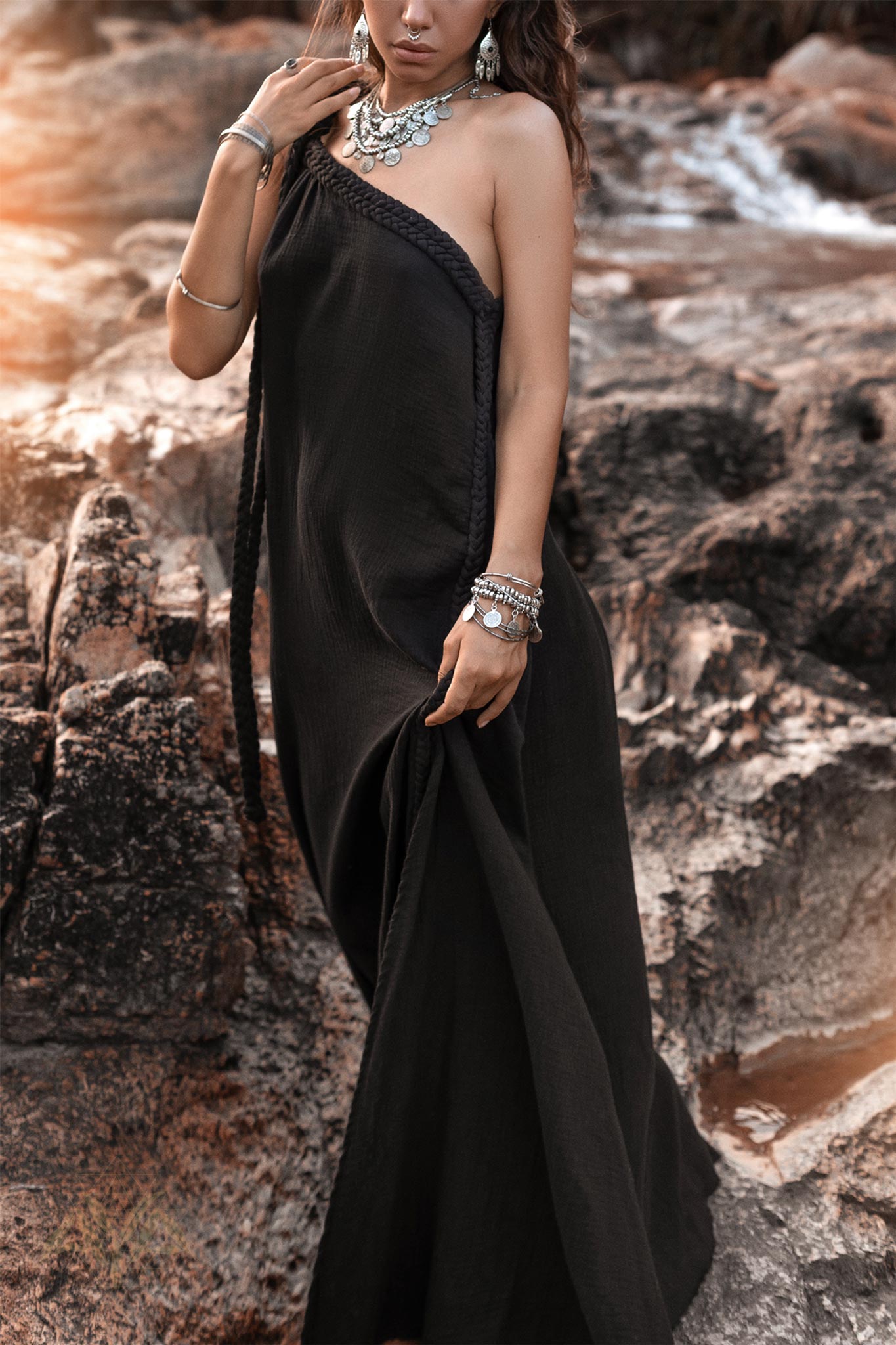 A woman stands on rocky terrain, wearing a cold shoulder Black Aurora Greek Goddess Dress by AYA Sacred Wear. She accessorizes with statement jewelry, including necklaces, bracelets, and rings, exuding a boho-chic aesthetic. The background features rugged rocks and a warm, glowing light.