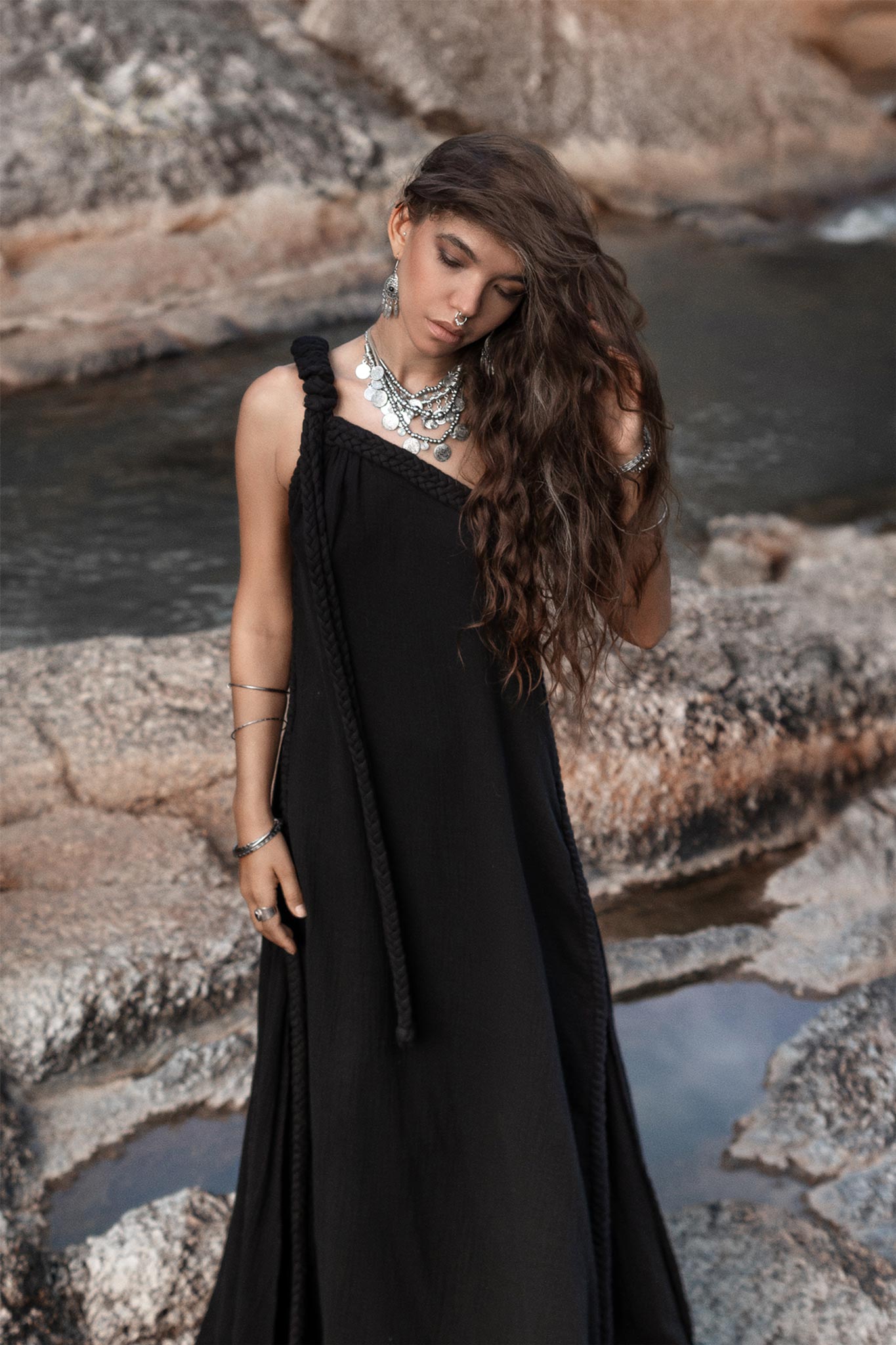 A woman with long, wavy hair and multiple necklaces stands outdoors on rocky terrain by the water. She is wearing a Black Aurora Greek Goddess Dress from AYA Sacred Wear, a cold shoulder maxi dress made of organic double-layer cotton. Her head is slightly tilted as she looks down with a serene expression, and her hand gently touches her hair.