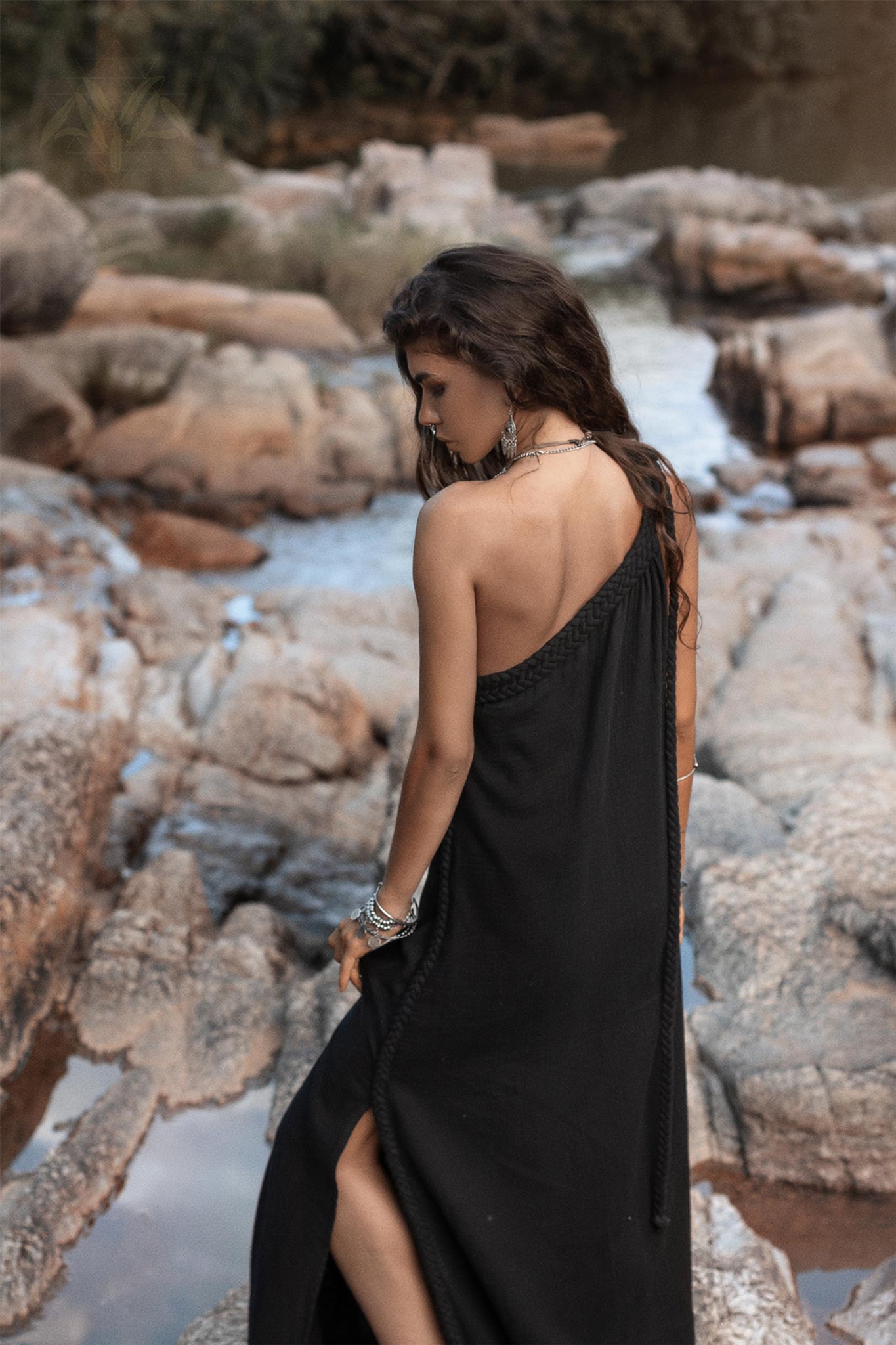 A woman with long wavy hair stands on rocky terrain near a stream, looking down. She is wearing a Black Aurora Greek Goddess Dress by AYA Sacred Wear and several bracelets. The background features rocks, water, and natural surroundings.
