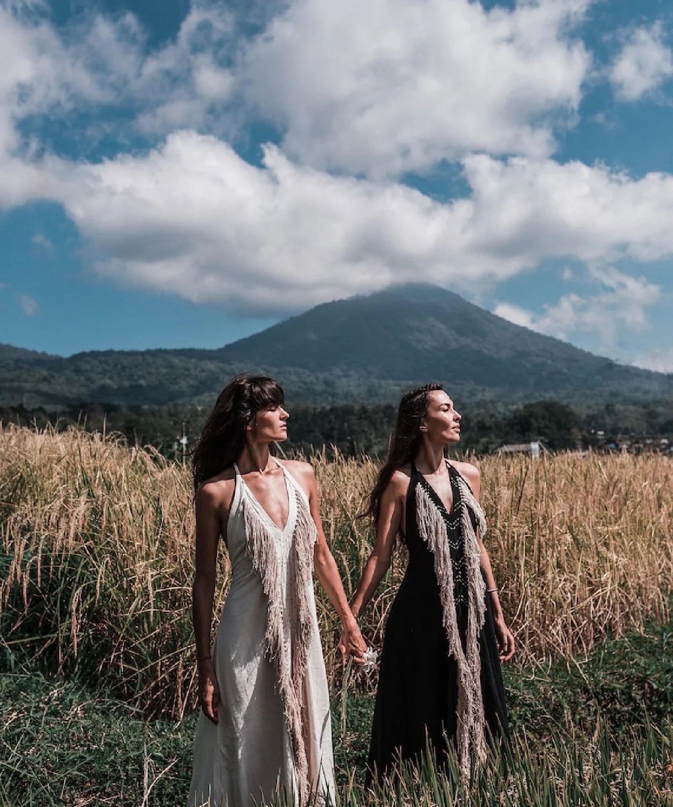 Two women wearing AYA Sacred Wear's Black Cotton Long Maxi Dress with open backs stand hand in hand in a grassy field, with mountains and a cloudy sky behind them.
