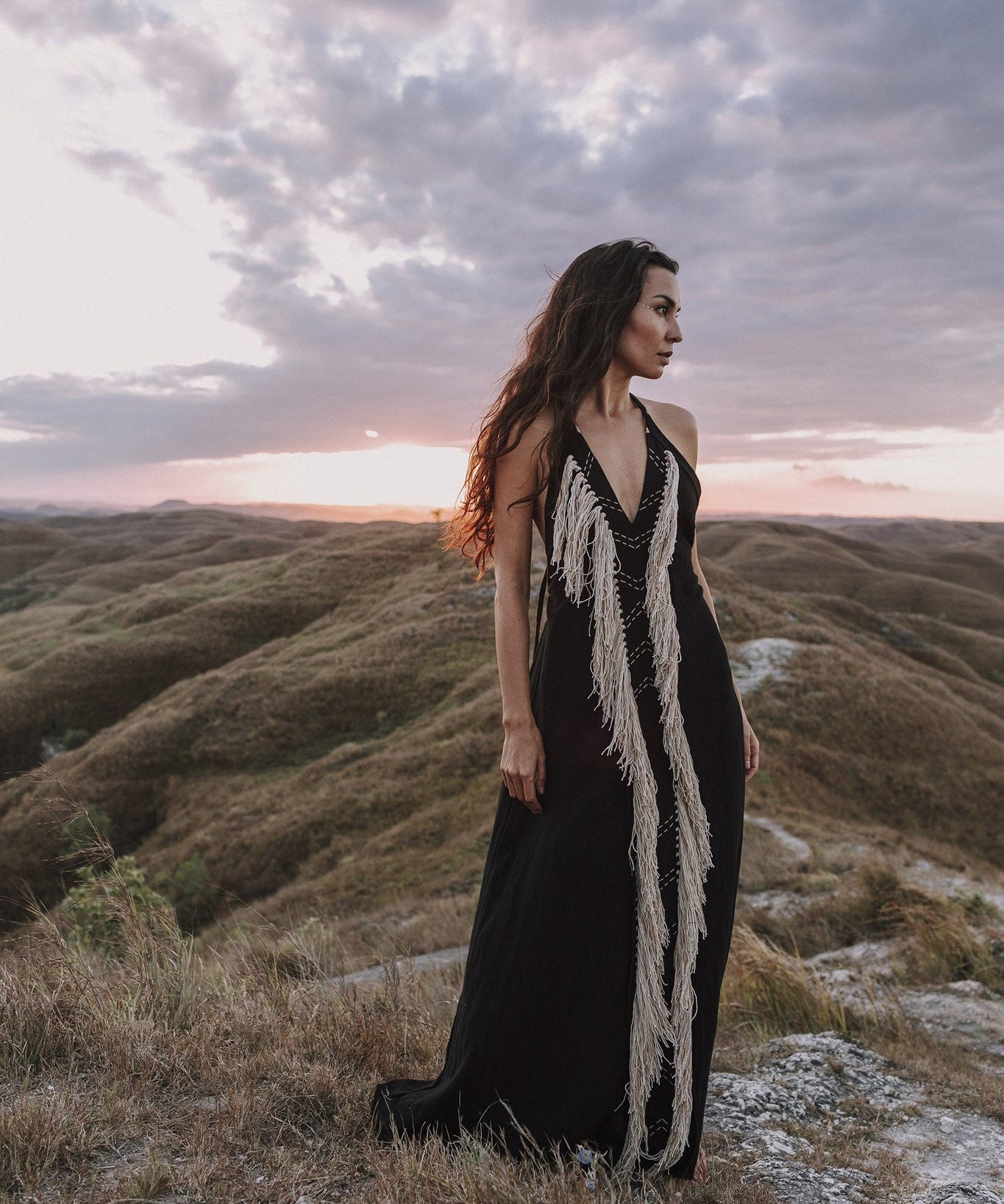 Clad in the Black Cotton Long Maxi Dress • Open Back Goddess Dress by AYA Sacred Wear, a woman stands gracefully on a grassy hilltop at sunset. She gazes into the distance, surrounded by rolling hills and a striking, cloudy sky in the backdrop.
