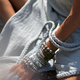 Close-up of a person wearing a light blue, textured Blue Aurora Greek Goddess Dress by AYA Sacred Wear, sitting with one hand resting on their knee. The hand and arm are decorated with numerous silver and metallic bracelets and a large ring. The soft, natural light suggests an outdoor setting.