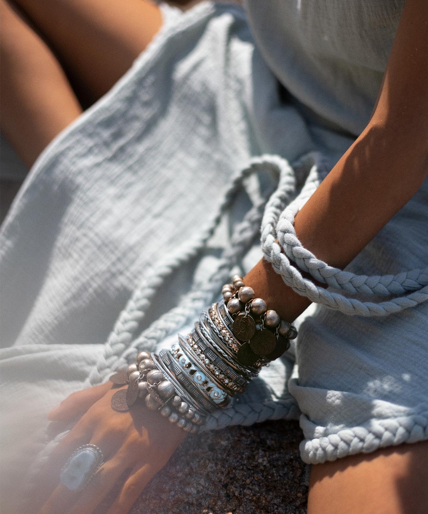 Close-up of a person wearing a light blue, textured Blue Aurora Greek Goddess Dress by AYA Sacred Wear, sitting with one hand resting on their knee. The hand and arm are decorated with numerous silver and metallic bracelets and a large ring. The soft, natural light suggests an outdoor setting.
