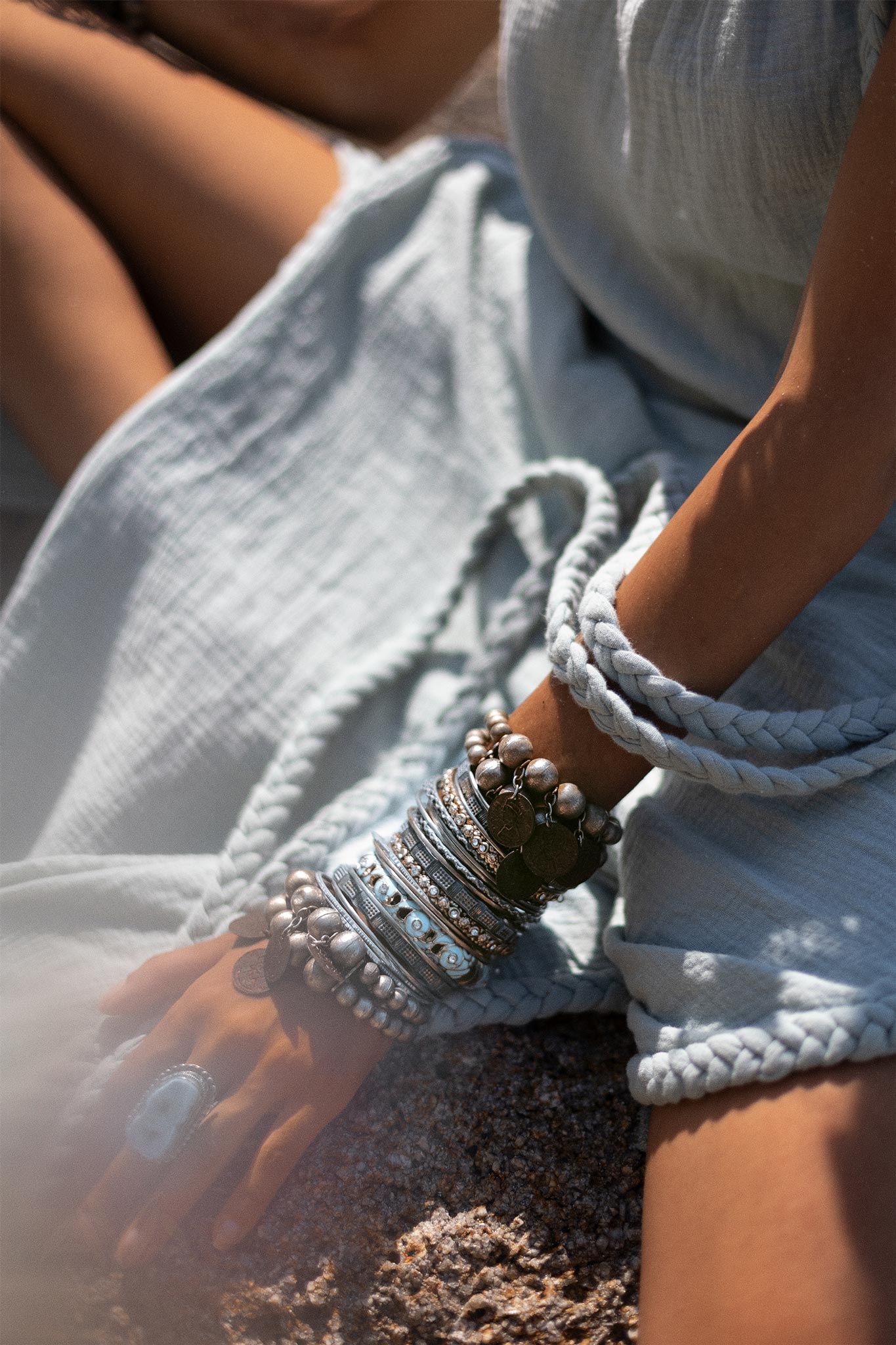 Close-up of a person wearing a light blue, textured Blue Aurora Greek Goddess Dress by AYA Sacred Wear, sitting with one hand resting on their knee. The hand and arm are decorated with numerous silver and metallic bracelets and a large ring. The soft, natural light suggests an outdoor setting.