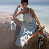 A person with dark hair is sitting on a large rock on a sandy beach, wearing an elegant Blue Aurora Greek Goddess Dress by AYA Sacred Wear. The Macrame Toga Dress, with its one-shoulder maxi design, is made of organic cotton. They are adorned with large, bold bracelets and earrings. The serene blue ocean and distant horizon provide a tranquil backdrop.