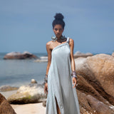 A woman stands confidently on a sandy beach surrounded by large rocks. She wears the light grey Blue Aurora Greek Goddess Dress by AYA Sacred Wear, featuring a one-shoulder macrame design with a slit, and large silver earrings. Her hair is styled in a high bun. The ocean and blue sky create a serene backdrop.
