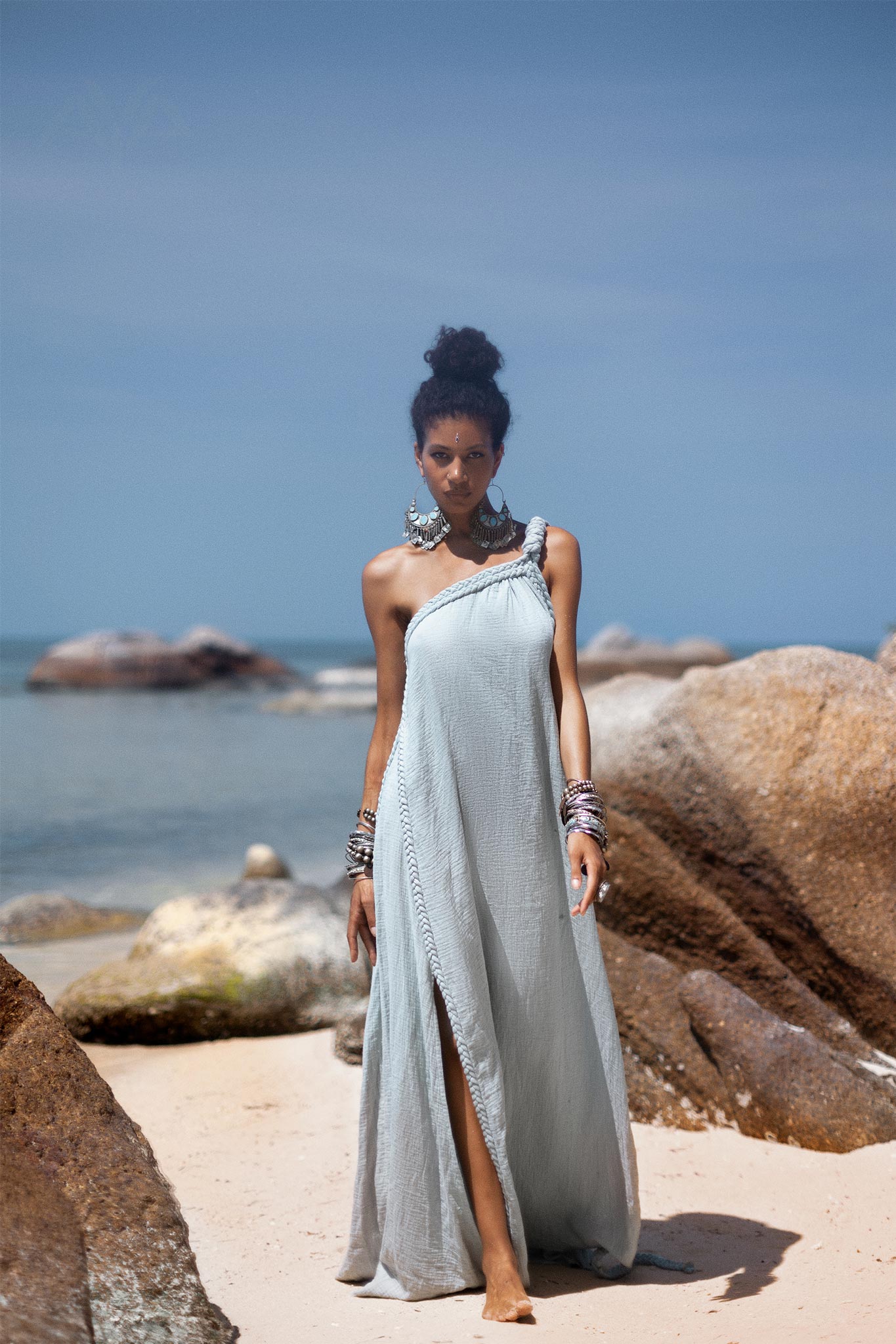A woman stands confidently on a sandy beach surrounded by large rocks. She wears the light grey Blue Aurora Greek Goddess Dress by AYA Sacred Wear, featuring a one-shoulder macrame design with a slit, and large silver earrings. Her hair is styled in a high bun. The ocean and blue sky create a serene backdrop.