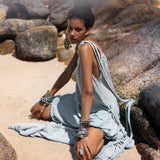 A woman with short, curly hair sits on a sandy beach surrounded by large rocks. She wears a flowing Blue Aurora Macrame Toga Dress, also known as the One Shoulder Maxi Dress from AYA Sacred Wear, adorned with multiple bracelets and earrings. She looks towards the camera with a soft, contemplative expression as the sun casts a warm glow on the serene scene.