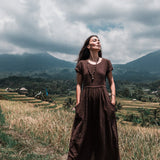 In a lush, grassy field with mountains looming in the background, a woman stands barefoot wearing an AYA Sacred Wear Bohemian Boho Dress for Women. The handwoven organic cotton fabric flows gracefully as she gazes upwards with a serene expression, embodying the essence of sustainable ethical fashion under a partly cloudy sky.