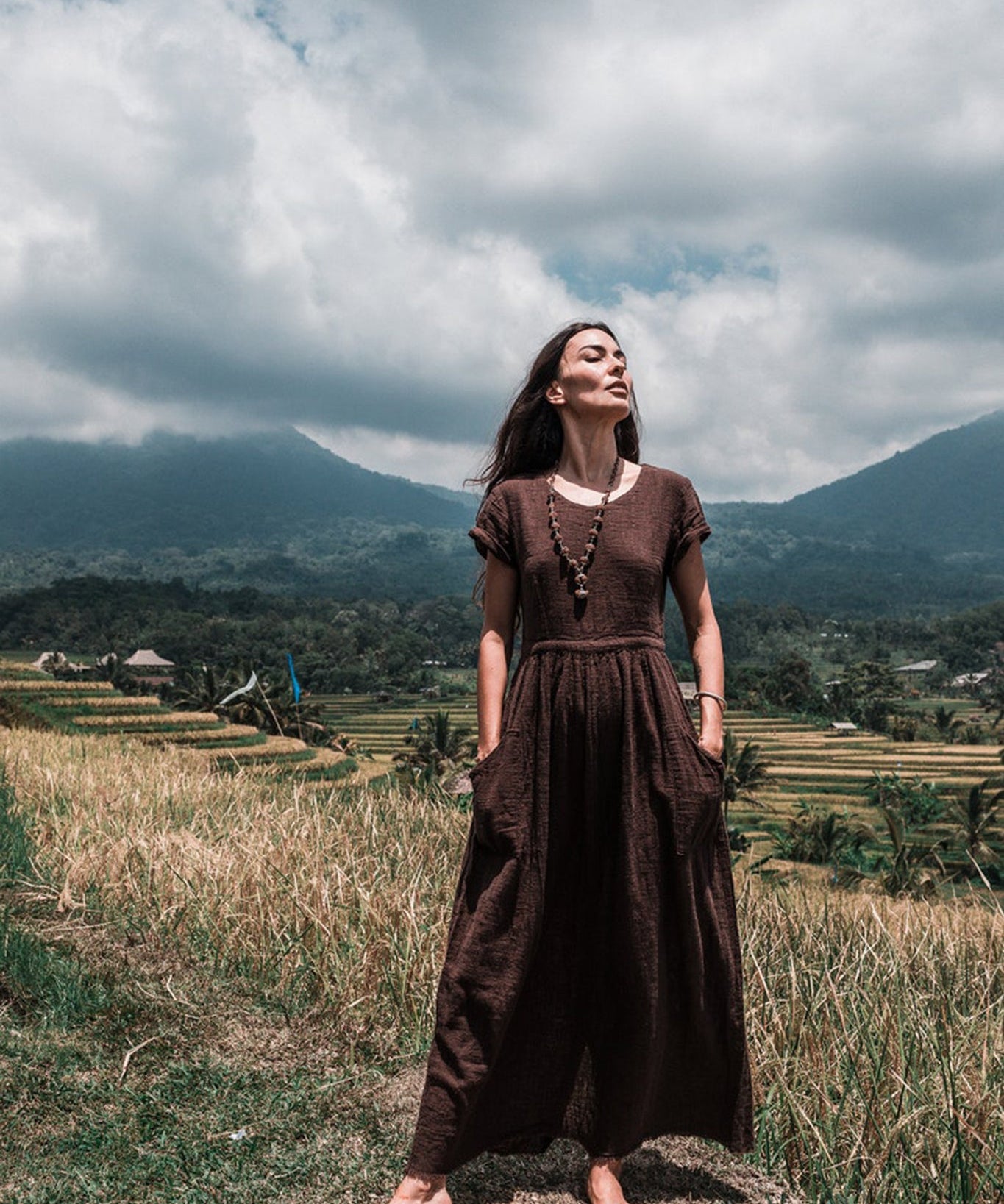 In a lush, grassy field with mountains looming in the background, a woman stands barefoot wearing an AYA Sacred Wear Bohemian Boho Dress for Women. The handwoven organic cotton fabric flows gracefully as she gazes upwards with a serene expression, embodying the essence of sustainable ethical fashion under a partly cloudy sky.