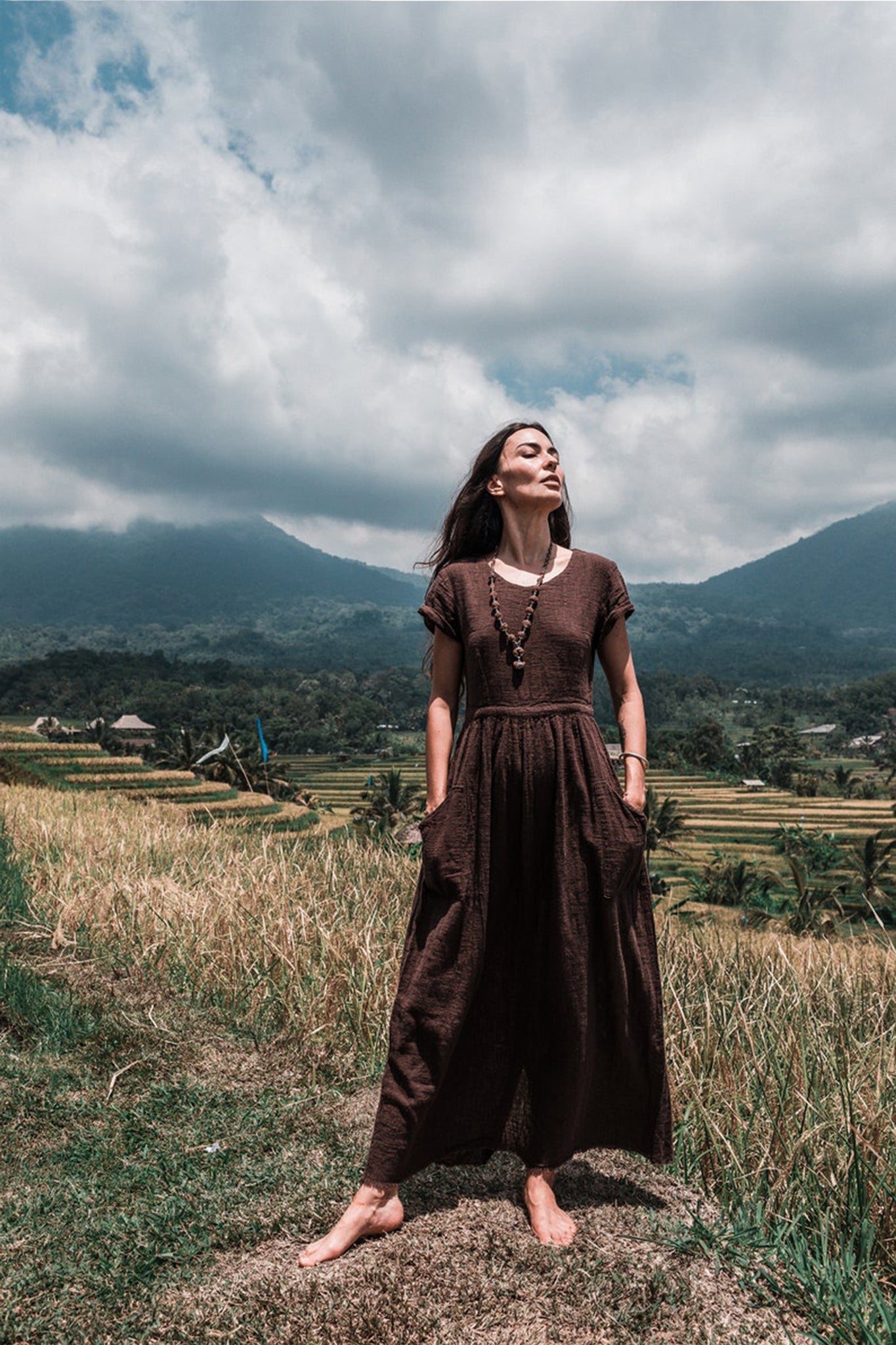 In a lush, grassy field with mountains looming in the background, a woman stands barefoot wearing an AYA Sacred Wear Bohemian Boho Dress for Women. The handwoven organic cotton fabric flows gracefully as she gazes upwards with a serene expression, embodying the essence of sustainable ethical fashion under a partly cloudy sky.