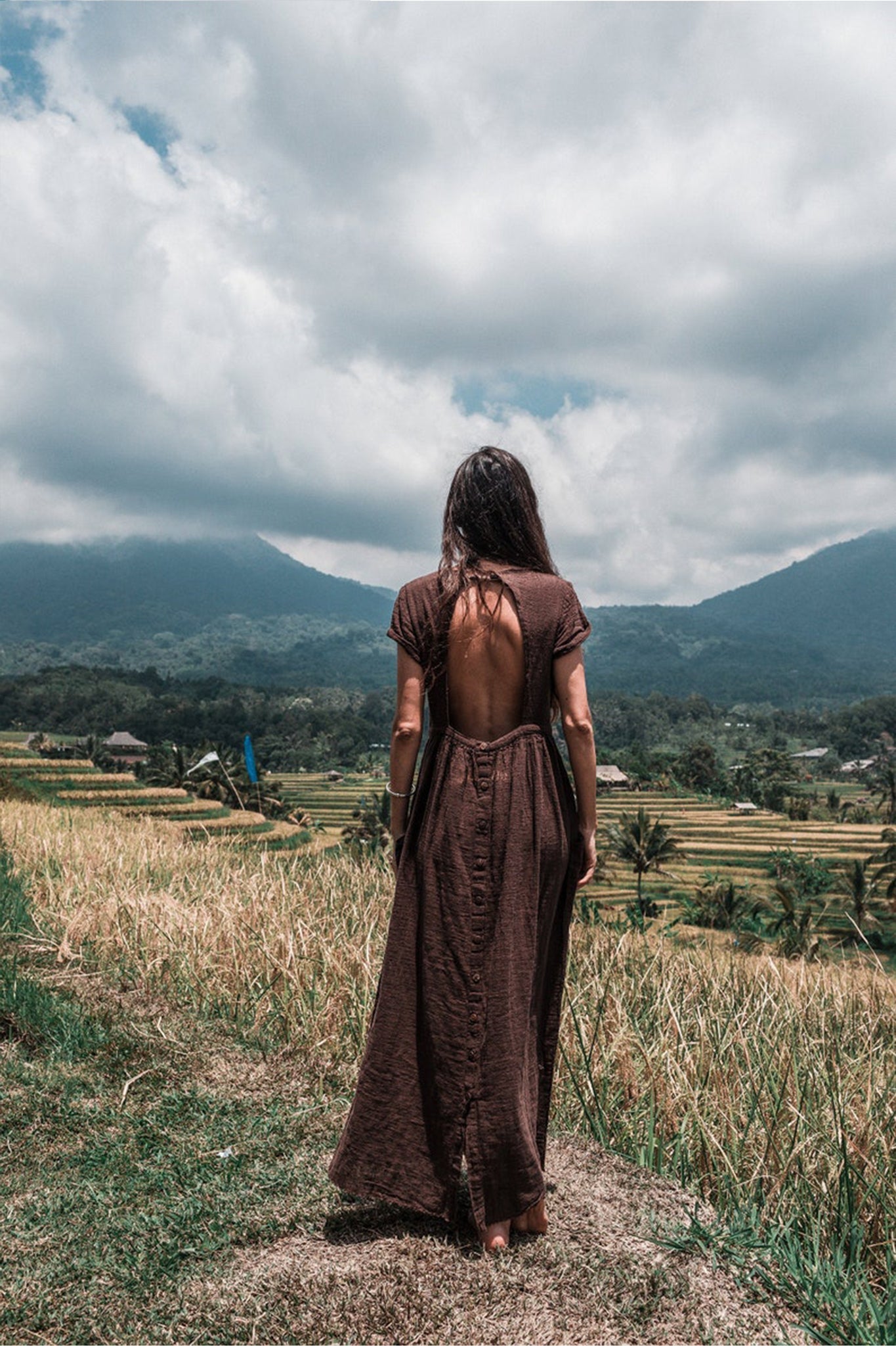 A woman stands on a grassy hill with her back to us, dressed in an AYA Sacred Wear Bohemian Boho Dress for Women. She gazes over verdant rice paddies and a mountainous view beneath a partly cloudy sky, truly representing the essence of sustainable ethical fashion.