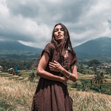 In a serene grassy field with mountains and cloudy skies as the backdrop, a woman showcases her style in a Bohemian Boho Dress for Women by AYA Sacred Wear. Made from handwoven raw cotton, this dress embodies her dedication to sustainable ethical fashion. Her long hair gently sways in the breeze, harmonizing perfectly with the tranquil natural environment.
