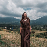 A woman wearing the Bohemian Boho Dress for Women by AYA Sacred Wear stands barefoot in a field, enveloped by lush green hills and mountains beneath a partly cloudy sky. She gazes upward with a serene expression, encapsulating the spirit of sustainable ethical fashion.