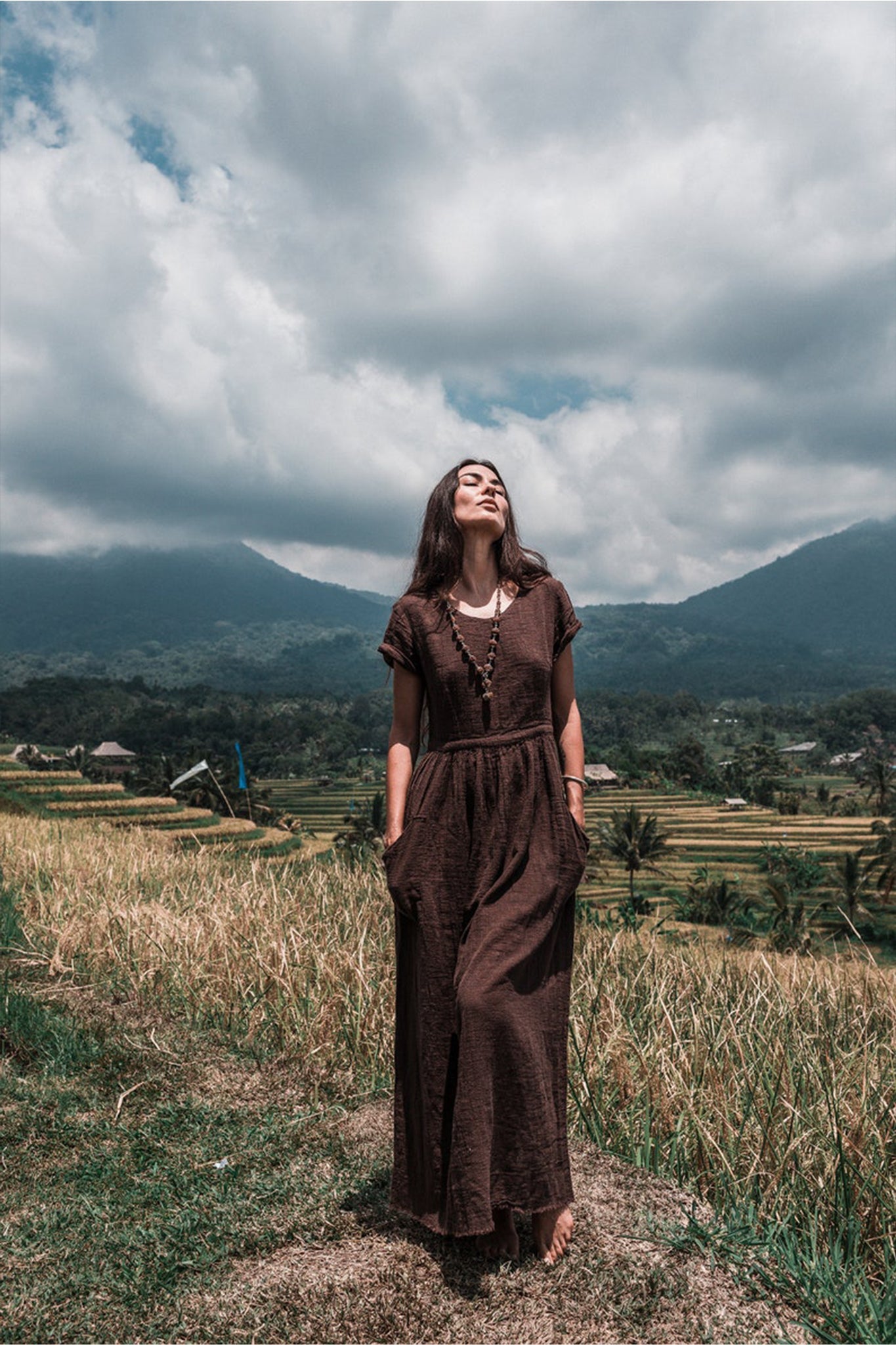 A woman wearing the Bohemian Boho Dress for Women by AYA Sacred Wear stands barefoot in a field, enveloped by lush green hills and mountains beneath a partly cloudy sky. She gazes upward with a serene expression, encapsulating the spirit of sustainable ethical fashion.
