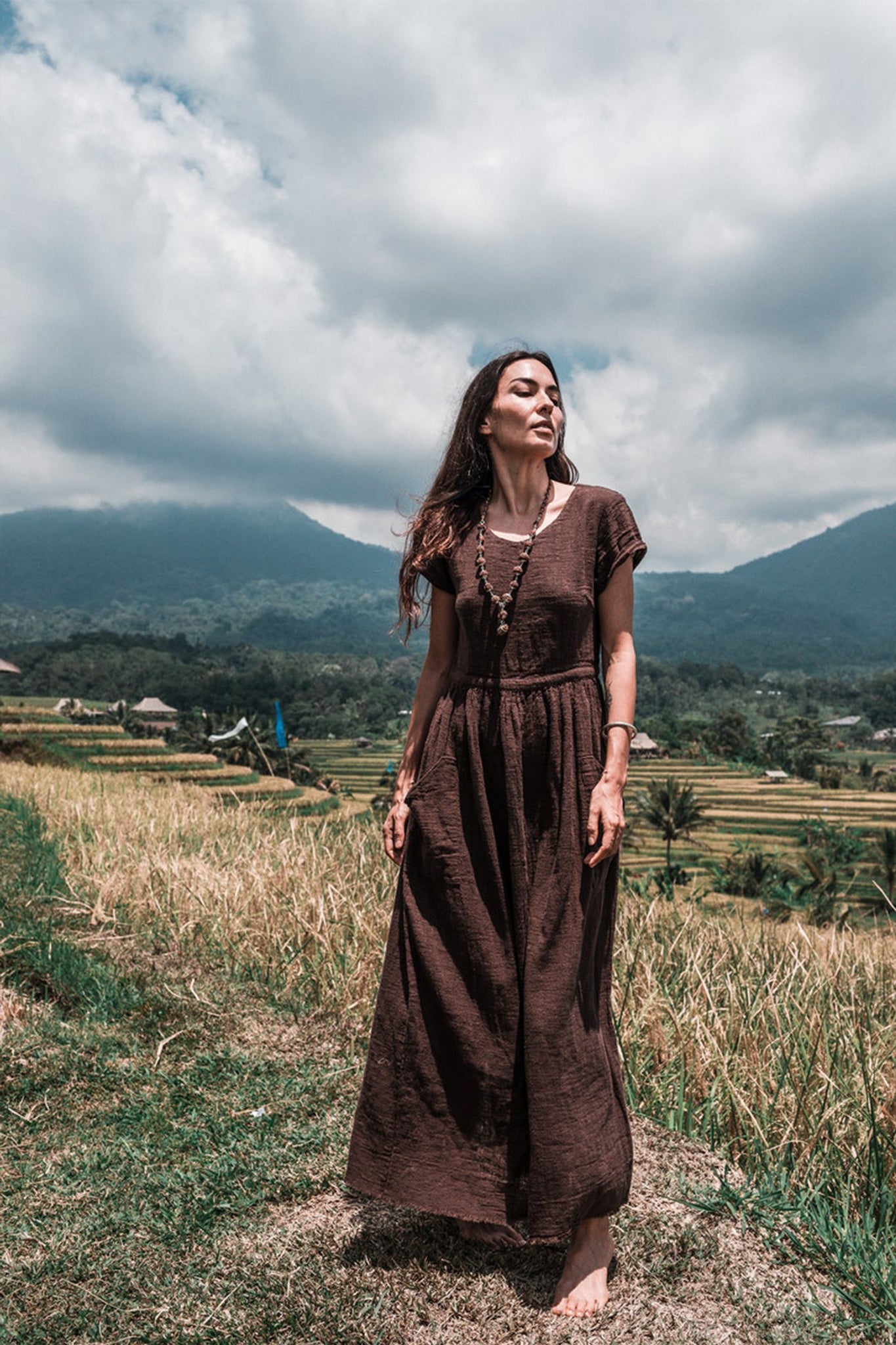 A woman wears an AYA Sacred Wear Bohemian Boho Dress as she stands barefoot on a grassy path, surrounded by lush green rice terraces and mountains beneath a cloudy sky.
