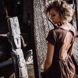 A woman with short hair wearing an AYA Sacred Wear Bohemian Boho Dress for Women stands alongside intricately carved wooden statues, her upper back displaying a tattoo. Natural light illuminates the detailed patterns on both the statues and her dress.