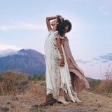 Two individuals stand back-to-back on rocky terrain, adorned in flowing outfits. One is wearing a Boho Beach Wedding Dress by AYA Sacred Wear, which has hand-loomed tassels and evokes a hippie wedding vibe. The other person wears a brown dress with Bohemian cutouts. In the background, a mountain rises against a pastel sky dotted with clouds, surrounded by brown grasses.