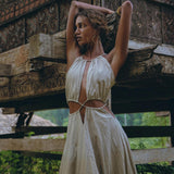 A person in a sleeveless, light-gray bohemian dress from AYA Sacred Wear stands with arms raised against a rustic, carved wooden structure. The Boho Bridesmaid Dress for Women, with its open back and cut-out sides, is crafted from organic fabric. Lush greenery can be seen in the background.