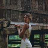 A person in an AYA Sacred Wear boho bridesmaid dress for women, featuring open back sides and a belly cutout, stands beside an ornately carved wooden structure with intricate patterns. The lush green foliage in the background complements the organic fabric and enhances the serene, natural ambiance.